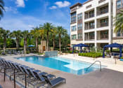 Thumbnail 25 of 35 - Pet-Friendly Apartments in Altamonte Springs FL - Nine12 Gateway - Sparkling Pool with Lounge Chairs, Navy Cabanas, Couches and Chairs, Landscaped Plants, Palm Trees, and View of Apartment Building