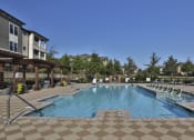 Thumbnail 26 of 36 - Greenville, SC Luxury Apartments - Velo Verdae - Resort-Style Swimming Pool with Sundeck, Submerged and Poolside Lounge Chairs, Seating Areas Under Pergolas, Checkered Tiles, and View of Apartment Building and Surrounding Trees
