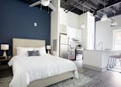 Thumbnail 7 of 25 - A bedroom with a large bed and a kitchen area in the background.