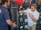 Thumbnail 13 of 31 - A man in a white t-shirt is punching a punching bag in a gym.