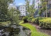 Thumbnail 3 of 35 - A pond in the middle of a grassy area with a building in the background.