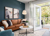 Thumbnail 6 of 21 - Apartments in Alexandria, KY, with In-Unit Washer and Dryer - Elevation Apartment Homes - Living Room with 9-Foot Ceiling, Wood-Style Flooring, Large Rug, Brown Couch, Blue Armchair, Round Center Table, Floor Lamp, 2 Large Wall Arts, Home Decor, Floor Length Curtains, and an Opened Doorway to the Balcony.