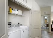 Thumbnail 23 of 38 - Luxury Apartments in Jacksonville, FL, with In-Unit Washer and Dryer - Terrabella - A Laundry Area with Washer, Dryer, Open Shelf, Wood-Style Flooring, and White Walls.