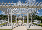 Thumbnail 43 of 67 - A white pergola structure is in the foreground of a park with benches and a building in the background.
