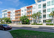 Thumbnail 23 of 25 - A red car is parked on the side of a road in front of a white building with orange accents.