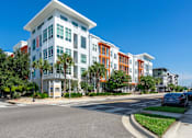 Thumbnail 24 of 25 - A modern multi-story building with a white and orange facade is situated on a street corner with cars parked along the curb.