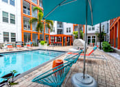 Thumbnail 19 of 25 - A blue umbrella is over a chair and table by a pool.