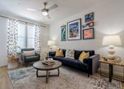 Thumbnail 5 of 25 - One-Bedroom Apartments in Tampa, FL - Havana Square - Living Room with Vinyl Plank Flooring, Area Rug, Sofa, Armchair, Coffee Table, Side Tables with Lamps, Wall Art, Ceiling Fan, and Large Window with Coverings.