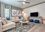 Thumbnail 17 of 29 - 2-Bedroom Luxury Apartments In Sarasota, FL - Botanic Waterside - Living Room With Wood-Finish Flooring, Area Rug, Sectional Couch, Coffee Table, Stools, Display Shelf, Wall-Mounted TV, Crown Molding, And Window.