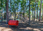 Thumbnail 23 of 30 - A red barrel sits in the middle of a forest with trees and buildings in the background.
