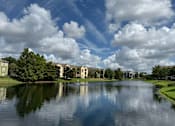 Thumbnail 28 of 31 - A serene lake surrounded by houses and trees under a blue sky with clouds.