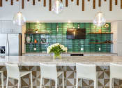 Thumbnail 22 of 23 - Daytona Beach, FL Apartments - Madison Pointe - Clubhouse Kitchen with Quartz Countertops, Green Tile Backsplash, Stainless Steel Appliances, Large Island with Seating, and Pendant Lights.