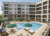 Thumbnail 1 of 16 - A large swimming pool surrounded by lounge chairs and umbrellas in front of a multi-story apartment building.