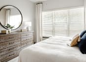 Thumbnail 28 of 34 - A bedroom with a white bed and a wooden dresser with a round mirror above it.