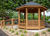 Thumbnail 20 of 28 - A wooden gazebo with a black roof is surrounded by a metal fence and a gravel area with plants.