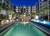 Thumbnail 37 of 39 - Orlando, FL, Pet-Friendly Luxury Apartments - NORA - Swimming Pool with a Sundeck, Fountain, Orange Lounge Chairs, Palm Trees, Warm Lights, and View of the Apartments.