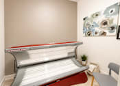 Thumbnail 15 of 31 - A tanning bed with a red and white cover is in a room with a painting on the wall.