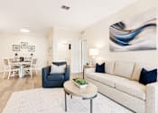 Thumbnail 3 of 31 - A living room with a white couch, a blue chair, a dining table with chairs, a lamp, and a painting on the wall.