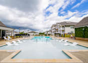 Thumbnail 26 of 40 - Luxury Apartments for Rent in Athens, GA - Classic City Flats - Resort-Style Saltwater Pool with Cabanas, In-Pool and Poolside Lounge Seats, Umbrellas, and View of Apartment Buildings in the Background.