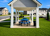 Thumbnail 59 of 65 - A pavilion with a white roof and grey walls is surrounded by a grassy area and a blue picnic table.