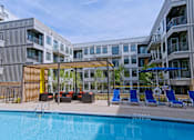 Thumbnail 12 of 32 - One Bedroom Apartments in Downtown Memphis, TN - The Rise - Poolside Outdoor Lounge Area with Brown and Red Wicker Style Table and Seating, Poolside Lounge Chairs, Umbrella, and Exterior View of the Property Building.