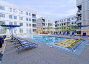 Thumbnail 11 of 32 - Studio Apartments in Downtown Memphis, TN - The Rise - Outdoor Resort-Style Pool with Surrounding Lounge Seating, Blue Umbrellas, Separate Lounge Areas with Wicker Tables and Seating, and an Exterior View of the Property Building.