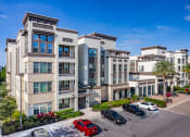 Thumbnail 1 of 35 - Luxury Apartments in Altamonte Springs, FL - Nine12 Gateway - Tan and White Property Exterior, with Street View of Parking, Landscaped Bushes, Plants, and Palm Trees