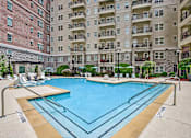 Thumbnail 2 of 39 - Atlanta, GA, Luxury Apartments Near Phipps Plaza - The Rocca - A Swimming Pool Surrounded by Lounge Chairs, Umbrellas, and a Multi-Story Apartment Building.