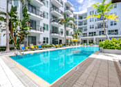 Thumbnail 1 of 22 - Tampa, FL Apartments Near Tampa Riverwalk - Society Westshore - Swimming Pool with Poolside Lounge Seats, Umbrella Tables and Chairs, Palm Trees, and View of Apartment Building.