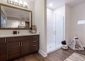 Thumbnail 11 of 22 - Studio Apartments in Tampa, FL - Society Westshore - Bathroom with Wood Plank Style Flooring, Large Vanity Mirror, Quart Countertop, Dark Wood-Tone Cabinetry, and Shower with Glass Door.