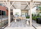 Thumbnail 27 of 30 - Pet-Friendly Luxury Apartments in Fort Worth, TX - The Union at River East - Grilling Station Underneath a Wooden Pergola, with Two Built-In Stainless Steel Grills, Tables, Armchairs, and a Trash Bin.