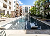 Thumbnail 1 of 30 - Two-Bedroom Luxury Apartments in Fort Worth, TX - The Union at River East - Swimming Pool with Small Fountains, In-Pool Loungers, Sun Loungers, Pergolas, Trees, and Metal Fence.