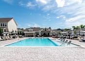 Thumbnail 53 of 56 - A swimming pool surrounded by a patio with chairs and a building in the background.
