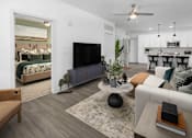 Thumbnail 16 of 50 - 2-Bedroom Apartments in Acworth, GA - The Brookstone - Living Room with Wood-Grain Plank Flooring, Area Rug, Sectional Couch, Side Table, Round Coffee Table, Ceiling Fan, Storage Cabinet, TV, and View of Bedroom and Kitchen.
