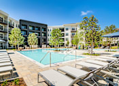 Thumbnail 24 of 26 - Pet-Friendly Luxury Apartments in Sugar Hill, GA - The Collier Sugar Hill - Large Resort-Style Heated Pool Surrounded by Loungers, Umbrellas, Cabanas, Trees, and Apartment Buildings.