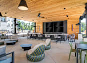 Thumbnail 17 of 26 - Sugar Hill, GA, Pet-Friendly Apartments - The Collier Sugar Hill - Spacious Outdoor Lounge with Wooden Ceiling, Recessed Lighting, Pendant Lamps, Ceiling Fan, Armchairs, Lounge Chairs, Low Coffee Tables, Square Dining Tables and Chairs, Speaker, Wall-Mounted Lighting, and a Bar Area with Wall-Mounted Screens, White Countertop, and Multiple Chairs.