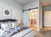 Thumbnail 8 of 28 - Pet-Friendly Apartments In Huntsville, AL - The Collins - Spacious Bedroom With Wood-Style Flooring, Area Rug, Bed, Nighstand with Lamp, High Ceilings, And A Large Closet With A Sliding Door
