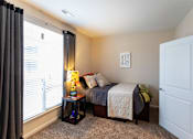 Thumbnail 10 of 25 - Dog-Friendly Apartments in Johnson City, TN - University Edge - Private Bedroom With Made Bed, Accent Pillows, Soft Carpet, Side Table Lamp, And Large Window With Blinds And Dark Curtains.