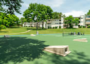 Thumbnail 25 of 35 - A green tennis court with a bench and a building in the background.