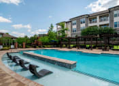 Thumbnail 23 of 36 - One-BR Apartments in Greenville, SC - Velo Verdae - Pool with Submerged Sun Lounge Chairs, Gazebos with Loungers, and Umbrella Table with Chairs