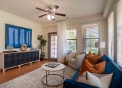 Thumbnail 14 of 36 - Pet Friendly Apartments in Greenville SC - Velo Verdae - Furnished Living Room with Natural Oak-Style Plank Flooring, Area Rug, Blue Sofa, White Armchair, Marble and Gold Round Coffee Table, Black and Tan TV Monitor, Standing Lamp, Large Windows with Blinds, Ceiling Fan, and House Plant in the Corner