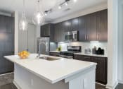Thumbnail 1 of 47 - Luxury Apartments Near Piedmont Park In Atlanta, GA - Venue Brookwood - Kitchen With Wood-Grain Flooring, Designer Finishes, Wood-Tone Flat-Panel Custom Cabinetry, Quartz Countertops, Stainless Steel Whirlpool Appliances, Built-in Microwaves, Designer Tile Backsplash With Under-Cabinet Lighting, Chrome Track Lighting, And Gourmet Kitchen Island with Pendant Lighting.