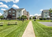 Thumbnail 14 of 15 - Pet-Friendly Student Apartments In Harrisonburg, VA - The Mill - An Outdoor Community With A Concrete Walking Path, Wooden Picnic Tables, And A Basketball Court.