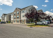 Thumbnail 1 of 15 - Four-Bedroom Student Apartments In Harrisonburg, VA - The Mill - An On-Site Parking Lot With Several Parked Cars, a Paved Roadway, Well-Maintained Green Lawns, and Landscaped Trees.