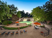 Thumbnail 13 of 32 - Columbus Apartments - Exterior View Of The Pool Area. The Area Has Lounge Chairs, Patio Sets With Umbrellas, Potted Plants, A Waterfall, Stairs, And Trees.