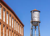 Thumbnail 31 of 33 - A water tower stands next to a brick building.