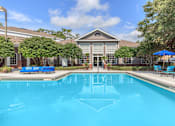 Thumbnail 22 of 36 - A swimming pool in front of a building with a blue umbrella and chairs.