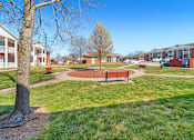 Thumbnail 2 of 14 - A park with a bench and a tree in the foreground and apartment buildings in the background.