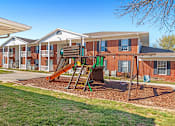 Thumbnail 3 of 14 - A playground with a slide and a green swing set in front of apartment buildings.