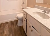 Thumbnail 12 of 35 - A white toilet sits in a bathroom with wood floors and a white sink.